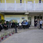 Več starejših ljudi sedi na klopci pred stavbo. Tri ženske stojijo in gledajo proti fotografu.