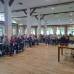 Večja skupina starejših ljudi sedi na stolih in opazuje nastopajočega starejšega gospoda.