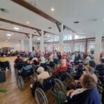 Večja skupina starejših ljudi sedi na stolih in opazuje nastopajočega starejšega gospoda.