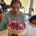 Starejša ženska piha svečko na torti, ki je na mizi.