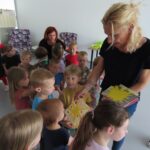 Skupina otrok in dve ženski. Desna ženska otrokom razdeluje papirnate izdelke.