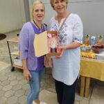 Dve ženski stojita, desna v rokah drži škatlo s steklenico.