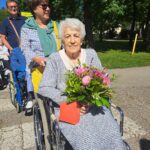 Ženska stoji za starejšo žensko, ki je na invalidskem vozičku in v rokah drži cvetje.