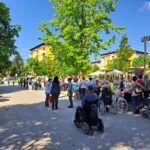 Več ljudi na invalidskih vozičkih s spremljevalci čaka na stranski cesti.