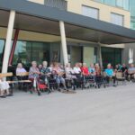 Več ljudi sedi pred stavbo, ena ženska pa stoji in govori v mikrofon.