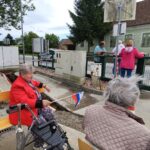 Dve starejši ženski sedita na klopcah ob cesti in v rokah držita manjše zastavice Slovenije.