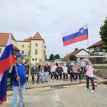 Več ljudi sedi na klopcah na prostem. Ženska in moški pa v rokah držita vsak svojo zastavo Slovenije.