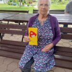 Starejša gospa sedi na stolu in v rokah drži papiranati izdelek.