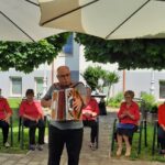Moški igra na harmoniko. V ozadju sedi 5 žensk v rdečih srajcah.
