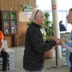 Dve ženski plešeta v paru. Starejši moški sedi na stolu iz palet in ju opazuje.