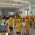 Več otrok stoji v rumenih majicah in kapah s šiltom pred skupino starejših ljudi.