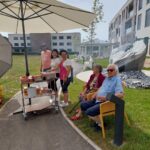 Dve ženski stojita zraven vozička z živili. Starejša moški in ženska pa sedita zraven na stolih.