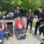 Več starejših ljudi sedi na klopcah. 4 mlajši ljudje in 1 deklica stojijo zraven starejše gospe na invalidskem vozičku.