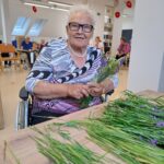 Starejša gospa sedi na invalidskem vozičku ob mizi na kateri je sivka.