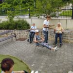 Več starejših ljudi sedi na stolih na prostem. Ženska jim streže kavo.