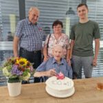 Štiri osebe ob mizi, na kateri je torta in lonec s cvetjem.