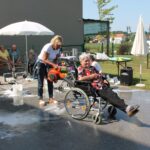 Ženska s pihalnikom piha v invalidskih voziček na katerem sedi starejša gospa.