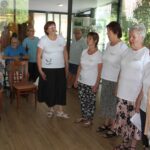 Istih šest žensk iz prve slike poje skupaj, tokrat stojijo na drugem delu sobe. Okoli njih sedi več ljudi, vključno s starejšimi moškimi in ženskami.