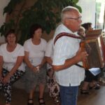 Moški igra harmoniko, medtem ko skupina žensk, nekatere sedijo, pojejo zraven. Ženske nosijo enake bele majice kot na prejšnjih slikah. V ozadju je še več starejših ljudi, ki spremljajo nastop.