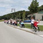 Skupina starejših ljudi v invalidskih vozičkih, ki se sprehajajo po pločniku. Spremljajo jih negovalci in prostovoljci. V ospredju je negovalka, ki dviguje roke v zrak, ob njej pa starejši gospod v rdeči majici, ki sedi v invalidskem vozičku in prav tako dviguje roke v zrak. V ozadju so drevesa in stavba z moderno arhitekturo.