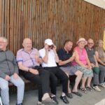 Skupina starejših ljudi sedi na klopi pred leseno steno. Vsak drži sladoled v rokah. Vsi so videti sproščeni in uživajo v prijetnem trenutku. Nekateri nosijo klobuke, drugi so oblečeni v lahka poletna oblačila.