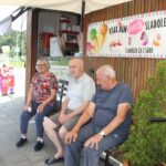 Trije starejši ljudje sedijo na klopi pred prodajalno sladoleda. Na zidu za njimi je plakat s sliko sladoleda in napisom "Vsak dan sveži sladoled". V ozadju je vidna prodajalna sladoleda z okenci in reklamo za sladoled. Vsi trije so nasmejani in videti zadovoljni.