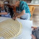 Starejša ženska z rezalnikom za testo reže trakove testa, ki so položeni na mizo. Ostale gospe, ki sedijo okoli mize, spremljajo postopek in pomagajo pri pripravi jedi.