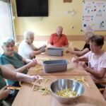Skupina starejših oseb sedi okoli mize v notranjem prostoru in čisti rumen fižol. Na mizi so posode, napolnjene s fižolom, ki ga pripravljajo za kuhanje. Vsi so osredotočeni na svoje delo, vzdušje pa je mirno in delovno.