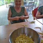 Starejša ženska s kratkimi sivimi lasmi sedi za mizo z velikim nasmehom, medtem ko čisti rumen fižol. Pred njo je velika kovinska skleda, napolnjena s fižolom, ozadje pa prikazuje zunanji vrt s pohištvom na terasi.