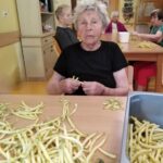 Starejša ženska v črni majici resno in osredotočeno čisti rumen fižol za mizo. Okoli nje so drugi stanovalci, ki sodelujejo pri istem opravilu, miza je polna fižola.