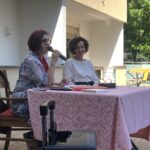 Bližnji posnetek dveh žensk, ki sedita za mizo s pink prtom, obe držita mikrofone. Ena ženska ima kratke kodraste lase in nosi belo bluzo, druga pa ima kratke rdeče lase, očala in je oblečena v pisano bluzo. Zdi se, da sta v pogovoru, na mizi pa so razprti knjige in papirji.
