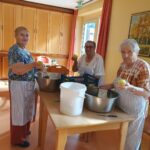 Tri starejše ženske, oblečene v predpasnike, stojijo okoli mize v toplo osvetljenem prostoru. Lupijo in pripravljajo jabolka, verjetno za recept, pri čemer uporabljajo različne kuhinjske pripomočke, kot so velike sklede in lupilci.