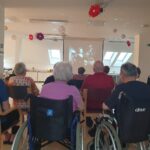 Skupina starejših oseb sedi v sobi in gleda črno-beli film, projiciran na zaslon. Nekateri so v invalidskih vozičkih, soba pa je okrašena z visečimi papirnatimi rožami in mehko osvetlitvijo.