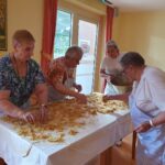 Štiri starejše ženske, vse oblečene v predpasnike, so zbrane okoli mize, prekrite z naribanim jabolčnim nadevom, verjetno za pecivo. Ena ženska drži velik lonec, ki verjetno vsebuje več nadeva, medtem ko ostale enakomerno razporejajo mešanico po mizi.