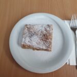 Bližnji posnetek kvadratnega kosa jabolčnega zavitka, posutega s sladkorjem v prahu, postreženega na belem krožniku. Vilica je postavljena na zložen prtiček poleg krožnika, pripravljena za uporabo.