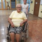 Starejša ženska v invalidskem vozičku, oblečena v rumeno majico in pisane hlače, se smehlja pred kamero. Za njo stoji mlado dekle, ki z nasmehom na obrazu potiska voziček. Dogodek poteka v svetli notranjosti z rumenimi stenami in steklenimi vrati v ozadju.