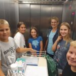 Skupina otrok in odraslih stoji v dvigalu, vsi se nasmejano gledajo v kamero. V dvigalu je voziček z medicinskimi pripomočki, vzdušje je sproščeno in prijateljsko.
