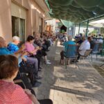 Skupina starejših oseb sedi na terasi, obrnjeni proti nastopajočim, ki se nahajajo izven kadra. Osebe so oblečene v barvita oblačila, vzdušje je sproščeno, terasa pa je pokrita z zelenim senčnikom, ki ustvarja prijetno senco.