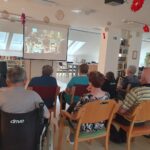 Skupina starejših oseb sedi v prostoru in gleda film, ki se predvaja na projekcijskem platnu. Nekaj oseb sedi na invalidskih vozičkih, drugi na stolih. Prostor je svetel, na stropu visijo dekoracije.