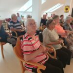 Več starejših oseb sedi v vrstah, osredotočeni na nekaj pred njimi (verjetno film). Nekateri imajo v rokah prigrizke. V ospredju sedi ženska v roza črtasti majici in z očali.