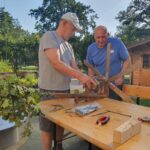 Dva starejša moška delata na leseni strukturi na prostem. Delata na vrtu, eden drži leseno kolo, drugi pa opazuje in pomaga.