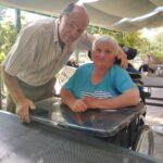 Slika prikazuje starejšega moškega, ki stoji poleg ženske na invalidskem vozičku. Oba sta nasmejana, medtem ko sta v senci pod nadstreškom na prostem. V ozadju so vidni drugi oskrbovanci in zelenje.