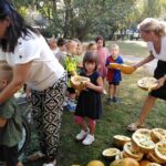 Skupina otrok in odraslih je zbrana na prostem med skupnostno dejavnostjo. Otroci držijo velike, na polovico razrezane buče, dve ženski pa jim pri tem pomagata. Dejavnost poteka na vrtu ali v parku, v ozadju so drevesa.