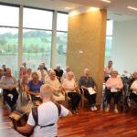 Skupina starejših ljudi sedi in gleda nastopajočega, ki igra harmoniko. Dogodek poteka v prostoru z velikimi okni, ki ponujajo razgled na podeželsko pokrajino, vključno z zelenimi hribi in drevesi. Starejši udeleženci so pozorni, nekateri držijo papirje, verjetno z besedili ali programom. Mnogi sedijo v invalidskih vozičkih, kar ustvarja topel in vključujoč ambient.