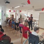 Skupina starejših ljudi sedi v svetli sobi in pozorno posluša moškega, ki igra harmoniko. Nekateri udeleženci so na invalidskih vozičkih, drugi pa sedijo na stolih, razporejenih v vrstah. Soba je okrašena z visečimi rdečimi in roza papirnatimi cvetovi, vzdušje pa je živahno.