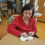 Starejša ženska v rdeči majici sedi za mizo in drži klobčič bele volne. V ozadju je prostor z rumeno-rjavo talno oblogo, invalidskim vozičkom in umivalnikom. Ženska je osredotočena na svoje delo.