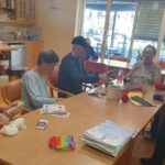 Skupina starejših ljudi sedi okoli mize in se ukvarja z ustvarjanjem z volno. Na mizi so različni materiali za ustvarjanje, kot so škarje, kartice in barvna volna. V ozadju je velika kuhinja z lesenimi elementi in oknom, skozi katero se vidi na vrt.