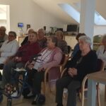 Skupina starejših ljudi sedi v osvetljeni sobi, urejeni s preprostim pohištvom, in pozorno gleda naprej. Nekateri udeleženci uporabljajo pripomočke za hojo ali invalidske vozičke. V ospredju je tudi miza z računalnikom, kjer je vidna filmska slika na zaslonu. Vzdušje je mirno in sproščeno.