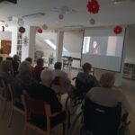 Starejši ljudje sedijo v vrstah in gledajo projekcijo na steno, kjer je prikazan film. Soba je nekoliko zatemnjena, kar poudarja osredotočenost udeležencev na projekcijo. Prostor je dekoriran z rdečimi papirnatimi cvetovi, ki visijo s stropa, in je videti udoben.