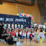 Otroci in vzgojiteljice v vrtcu praznujejo, obkroženi z barvitimi baloni in odrom, okrašenim s sliko otrok. Napisa na ozadju: 'Naš vrtec praznuje'.